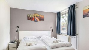 Una cama doble con ropa blanca, una mesita de noche con lámpara y una ventana con cortinas.