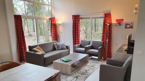 Una sala de estar moderna con sofá, sillón y mesa de centro, ventanas amplias con cortinas y vistas a árboles.