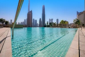 Une piscine à l’eau d’un bleu cristallin, entourée d’une terrasse carrelée. Au fond, on aperçoit des immeubles de grande hauteur, dont un très grand gratte-ciel.