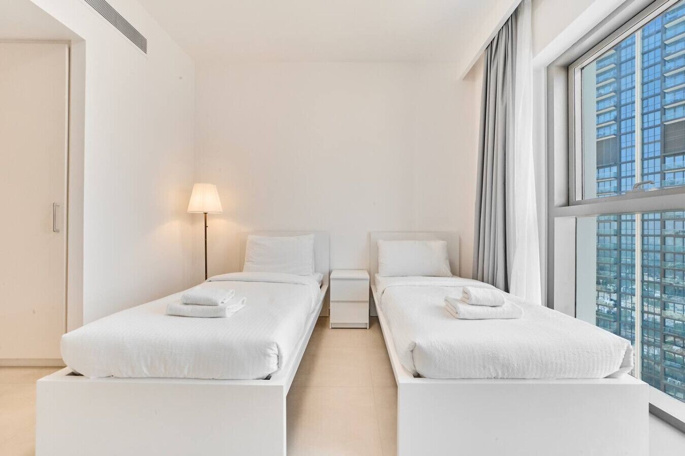 Quarto de hotel moderno com duas camas de solteiro, mesa de cabeceira com abajur e uma janela grande com vista para a cidade.