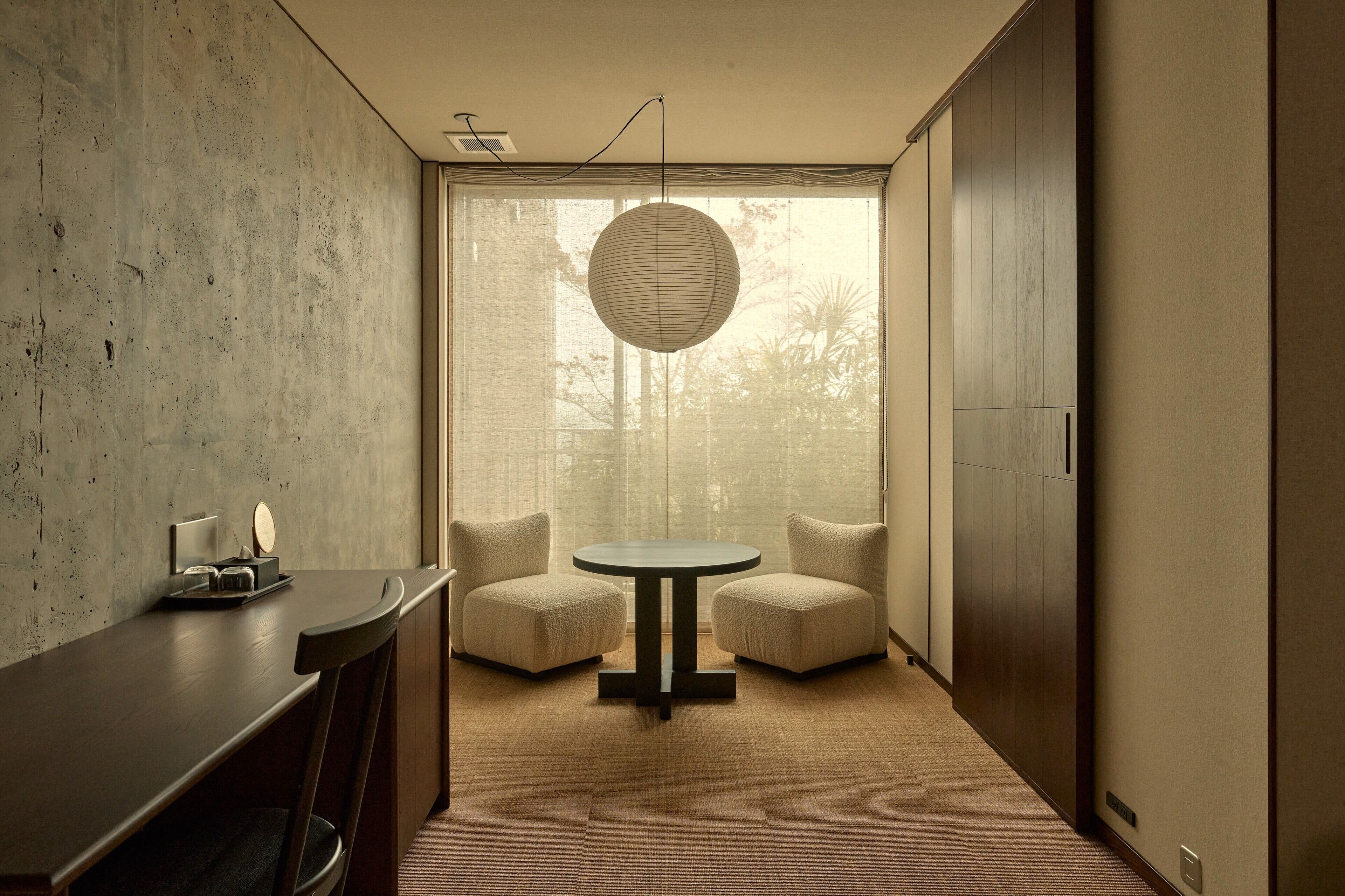 A modern room with a round table, two chairs, a concrete wall, and a large window with a view of trees.