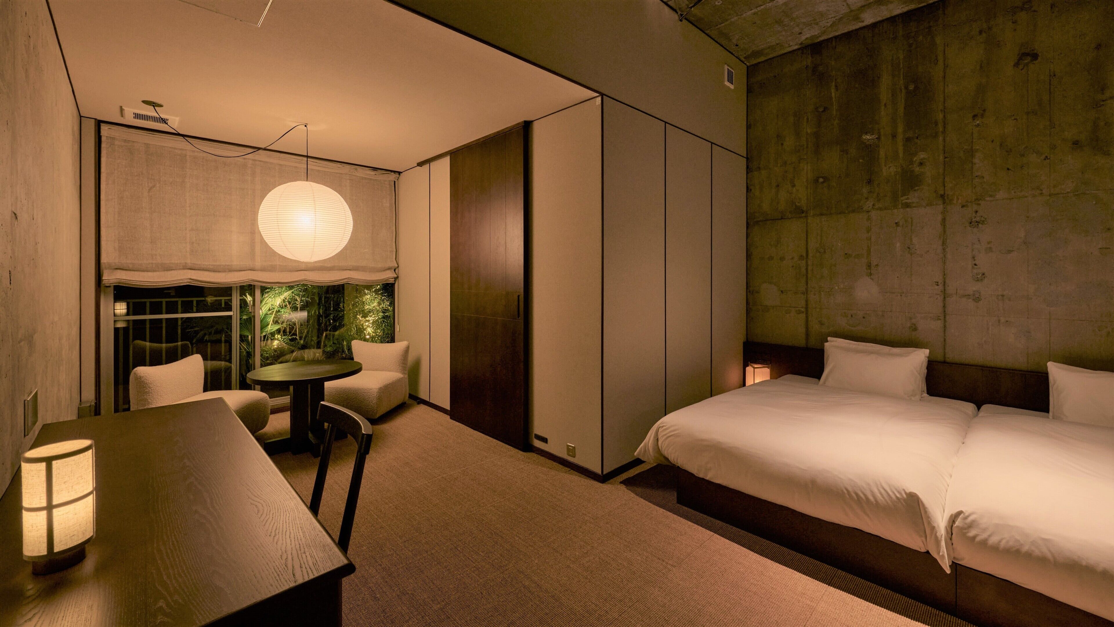 A modern hotel room with a bed, a small table, a chair, a lamp, and a view of greenery through a window.