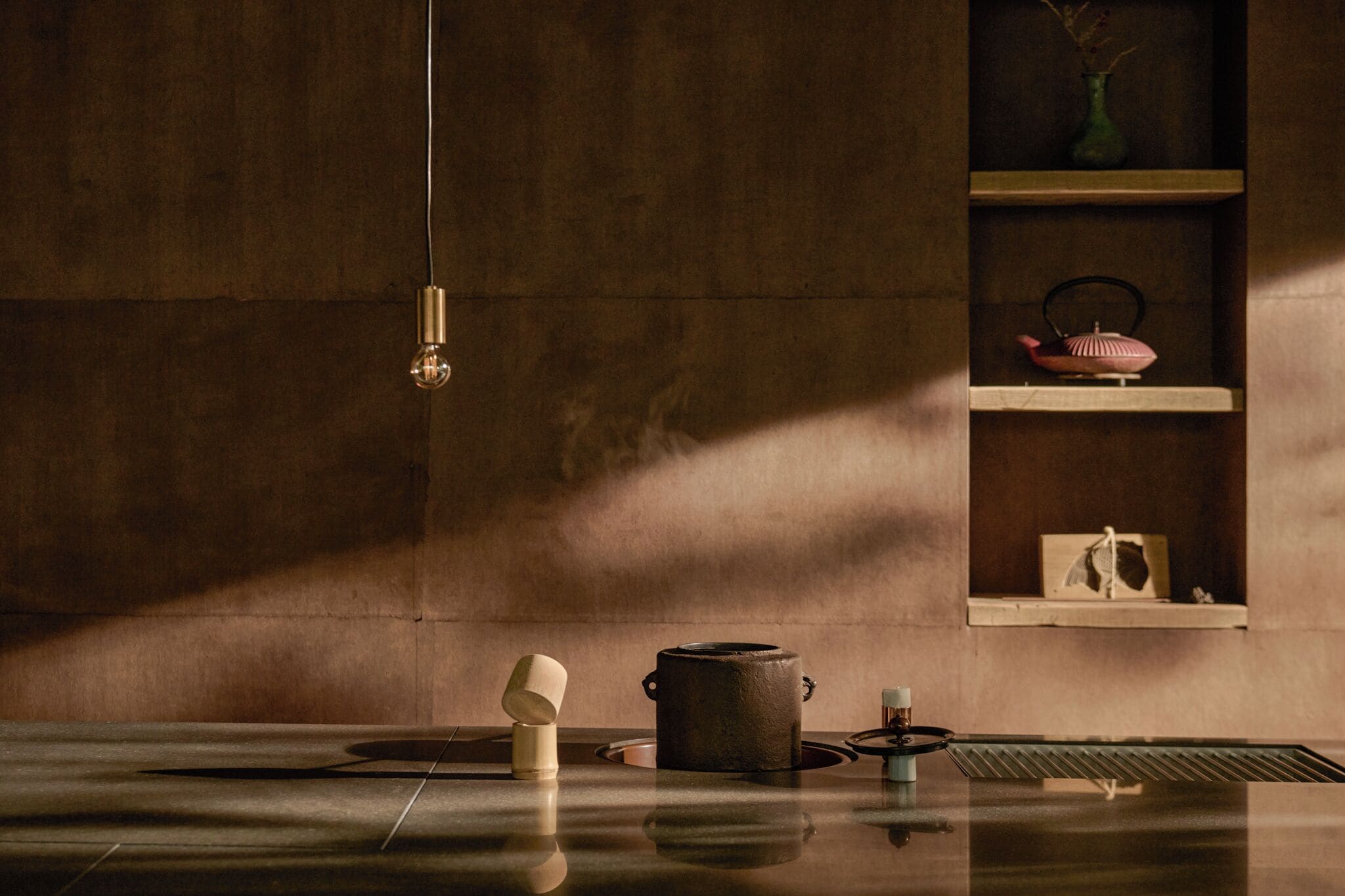 A minimalist kitchen with a hanging light, a pot, a teapot, and a shelf with decorative items.
