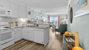 Una cocina moderna con armarios blancos, horno empotrado y lavavajillas. Cuenta con una zona de comedor con una silla verde y una mesa, y vistas al exterior a través de una ventana.