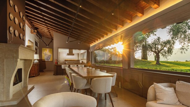 Une salle à manger spacieuse avec une longue table en bois et des chaises blanches, une cheminée et une grande fenêtre donnant sur un paysage verdoyant.
