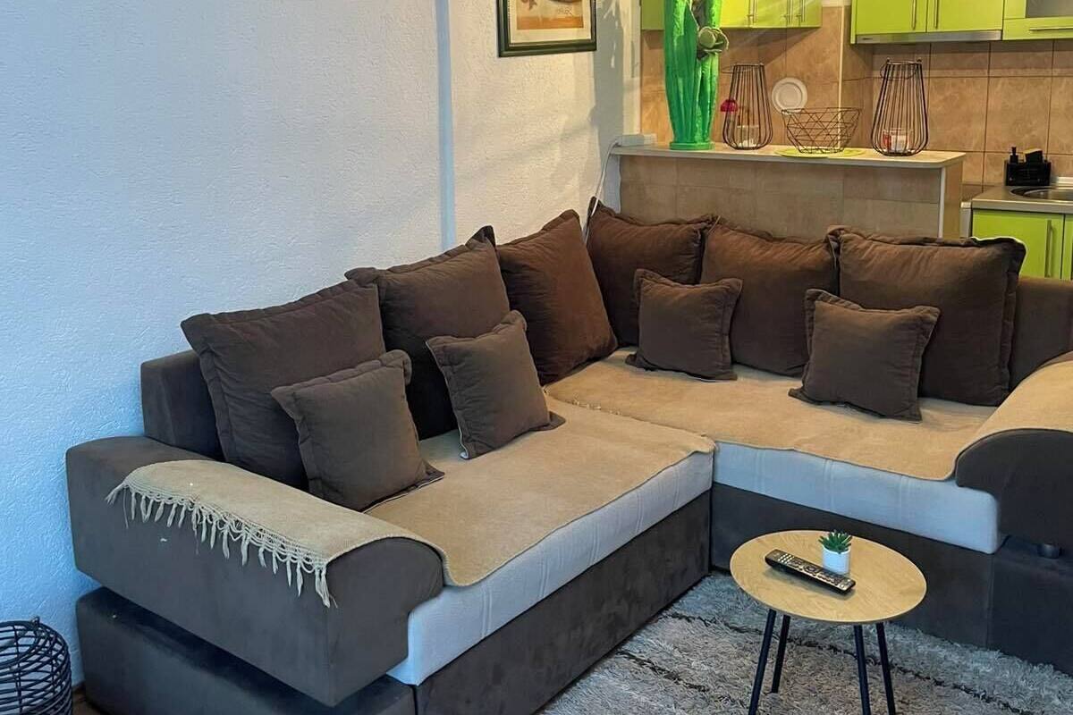 A living room with a sectional sofa, a small round table with a remote control, and a green kitchen in the background.