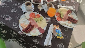 Una mesa dispuesta al aire libre con diversos alimentos para el desayuno, como pan, huevos, carnes y verduras, además de bebidas y salsas.