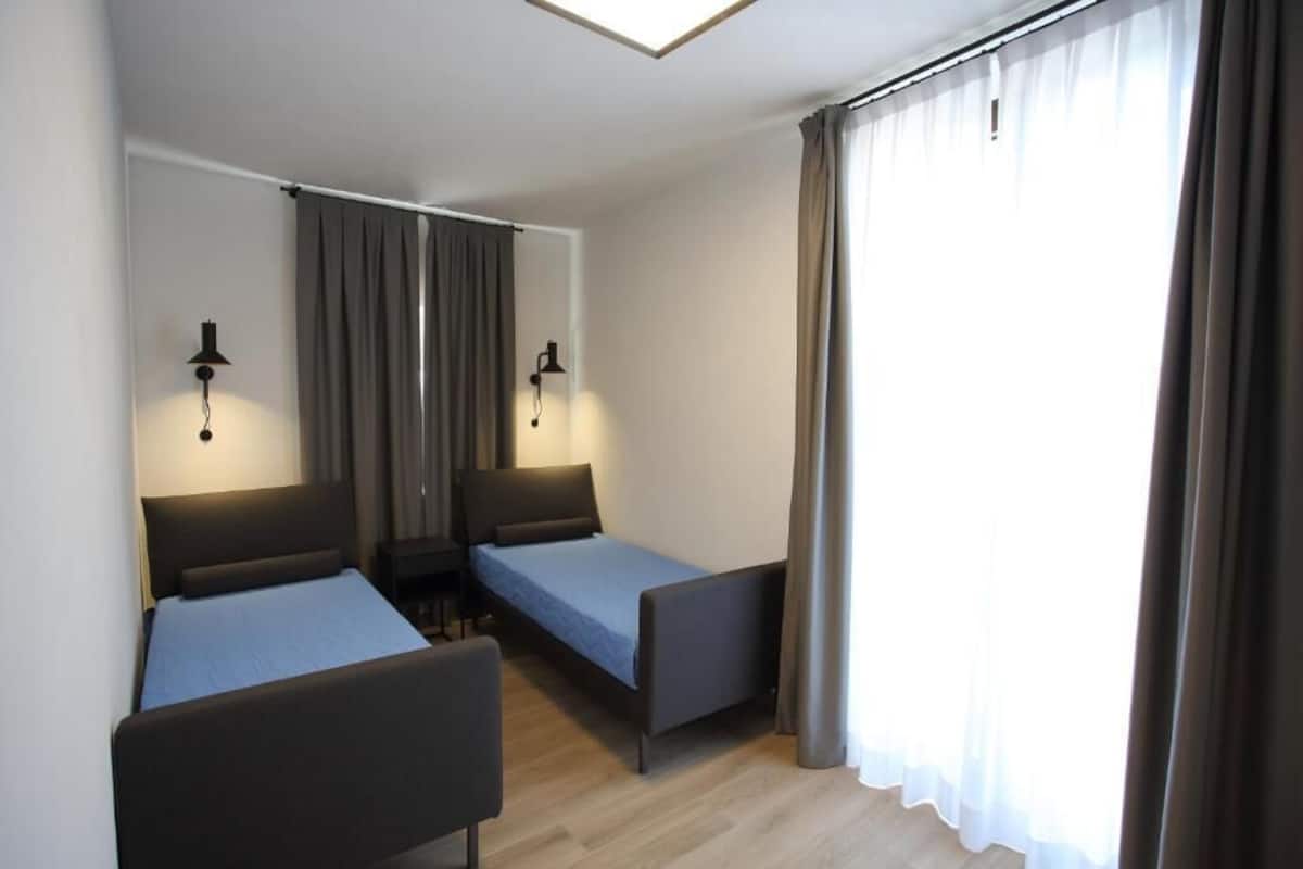 A room with two single beds, a desk, and a chair. There are two wall-mounted lamps.