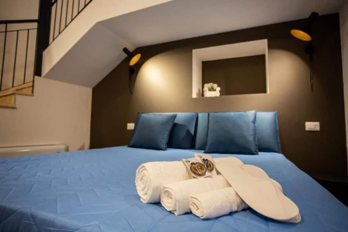 A hotel room with a blue bedspread, a neatly made bed with rolled white towels and a pair of slippers, a small white vase on the nightstand, and a dark wall with a light fixture.