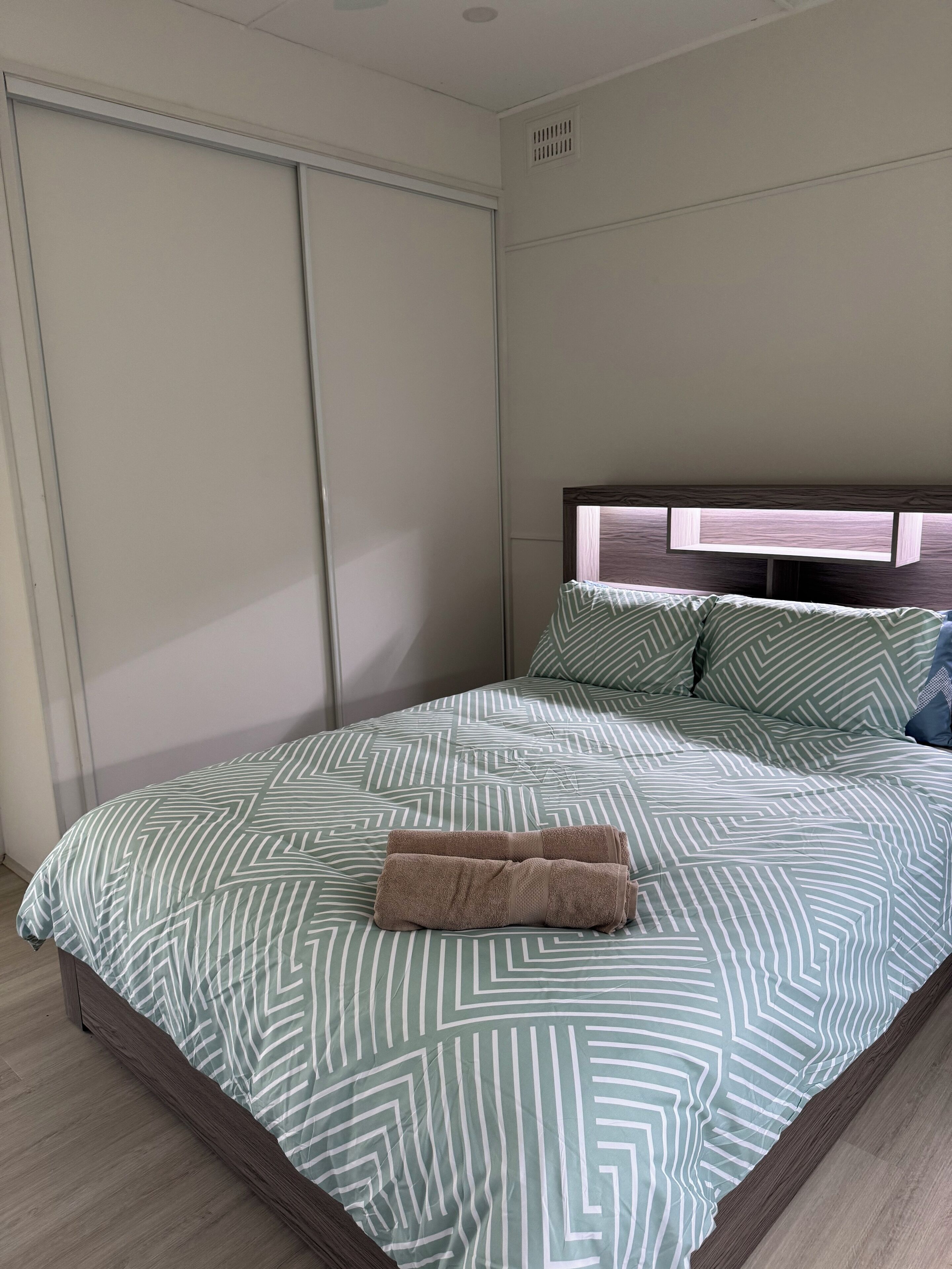A neatly made bed with a patterned bedspread, a folded towel, and a headboard with a light wood finish.
