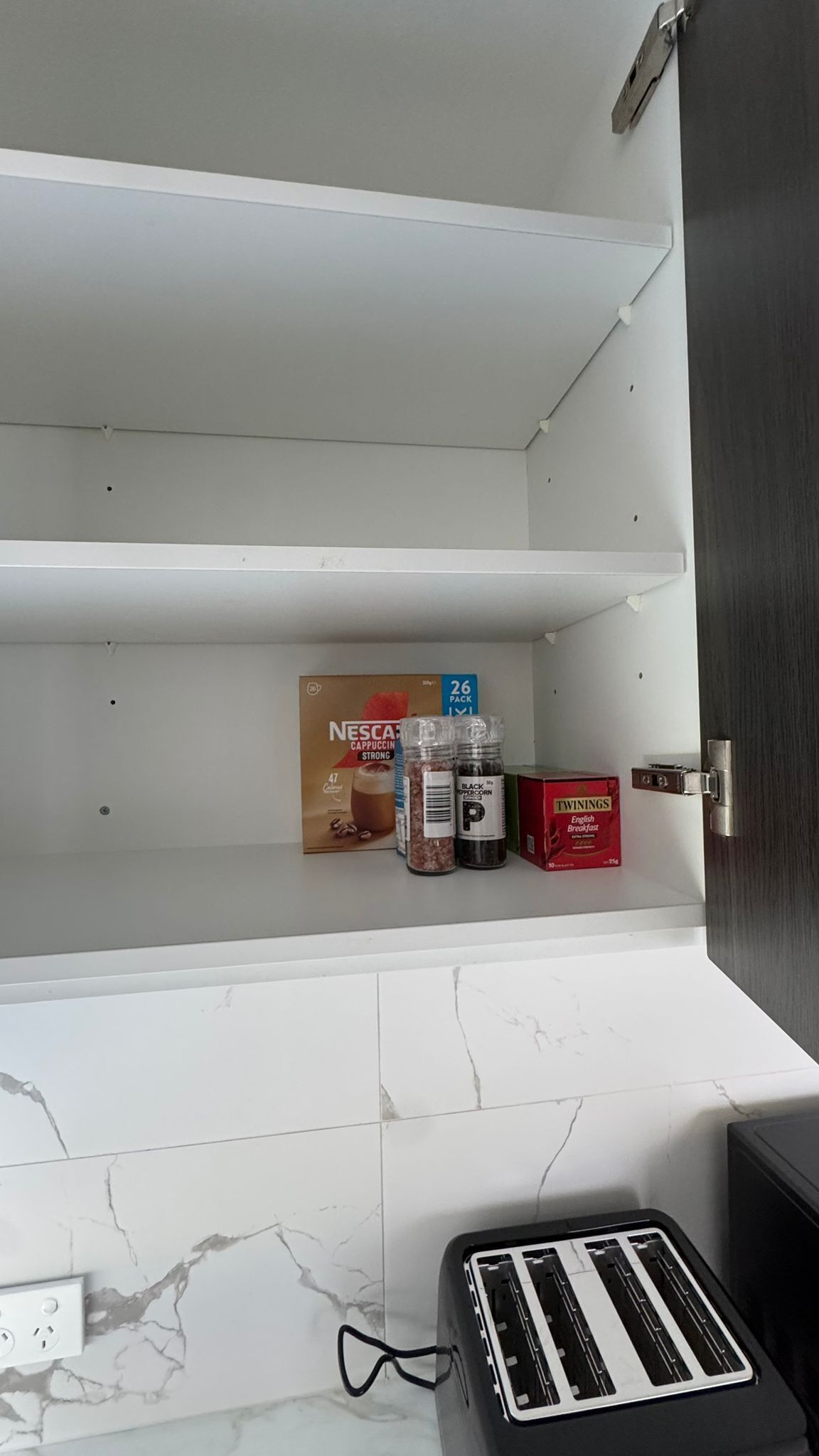 A kitchen with a white tiled backsplash, a toaster, and a shelf with a Nescafé cappuccino pack, black pepper, and Twinings English Breakfast tea.