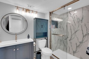 A modern bathroom with a white toilet, a blue cabinet, a round mirror, and a glass shower enclosure. - 2B/2B Marina Towers 704, Bay View, Heated Pool! Hot tub! (Fort Myers Beach)