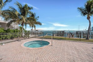A swimming pool with a metal ladder, surrounded by a brick patio and palm trees, with a view of the ocean and a fence. - 2B/2B Marina Towers 704, Bay View, Heated Pool! Hot tub! (Fort Myers Beach)