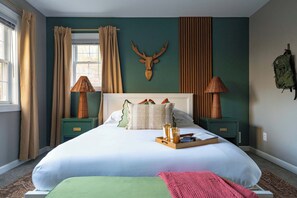 Une chambre avec un grand lit, une décoration murale représentant une tête de cerf en bois, deux tables de chevet avec des lampes et un sac à dos accroché au mur.