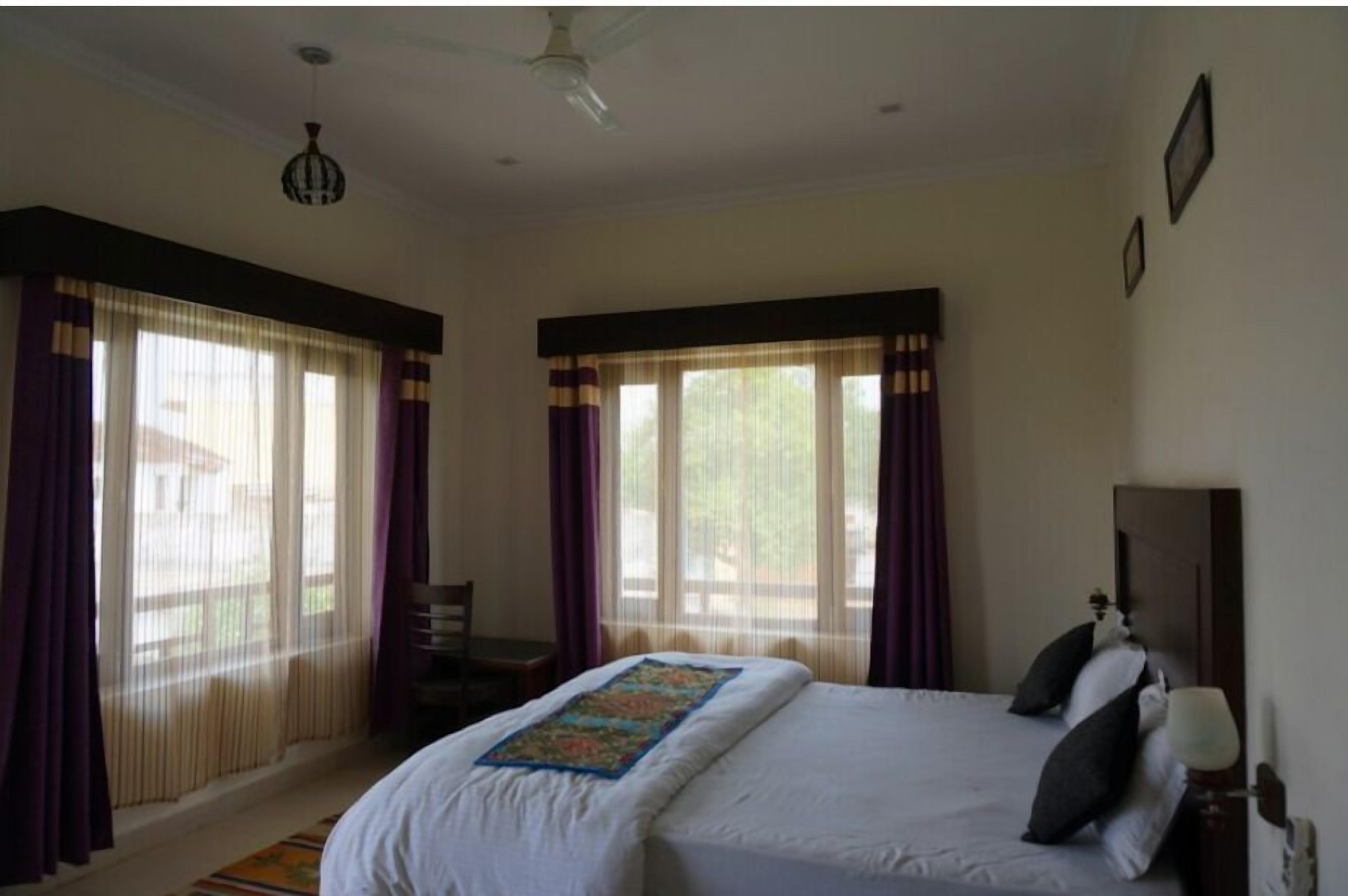 A hotel room with a bed, a desk, and a chair. There are curtains on the windows, and a ceiling fan is visible.