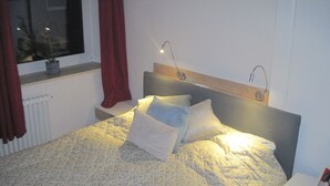 A neatly made bed with a quilted headboard, two bedside lamps, and a window with red curtains.