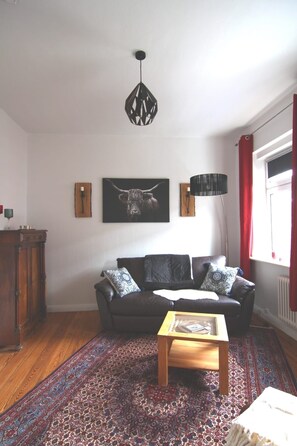 Ein Wohnzimmer mit einem dunkelbraunen Sofa, einem gemusterten Teppich, einem Holztisch und einer schwarzen Pendelleuchte, sowie einem gerahmten Kunstwerk mit einem Stierkopf.