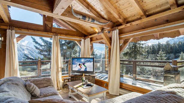 Un accogliente chalet in legno con un grande schermo televisivo che mostra una scena di sci, posti a sedere confortevoli e vista sulle montagne innevate.