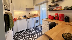 Eine kleine Küche mit weißen Schränken, einer Waschmaschine, einer Kaffeemaschine und einem Regal mit Pflanzen.