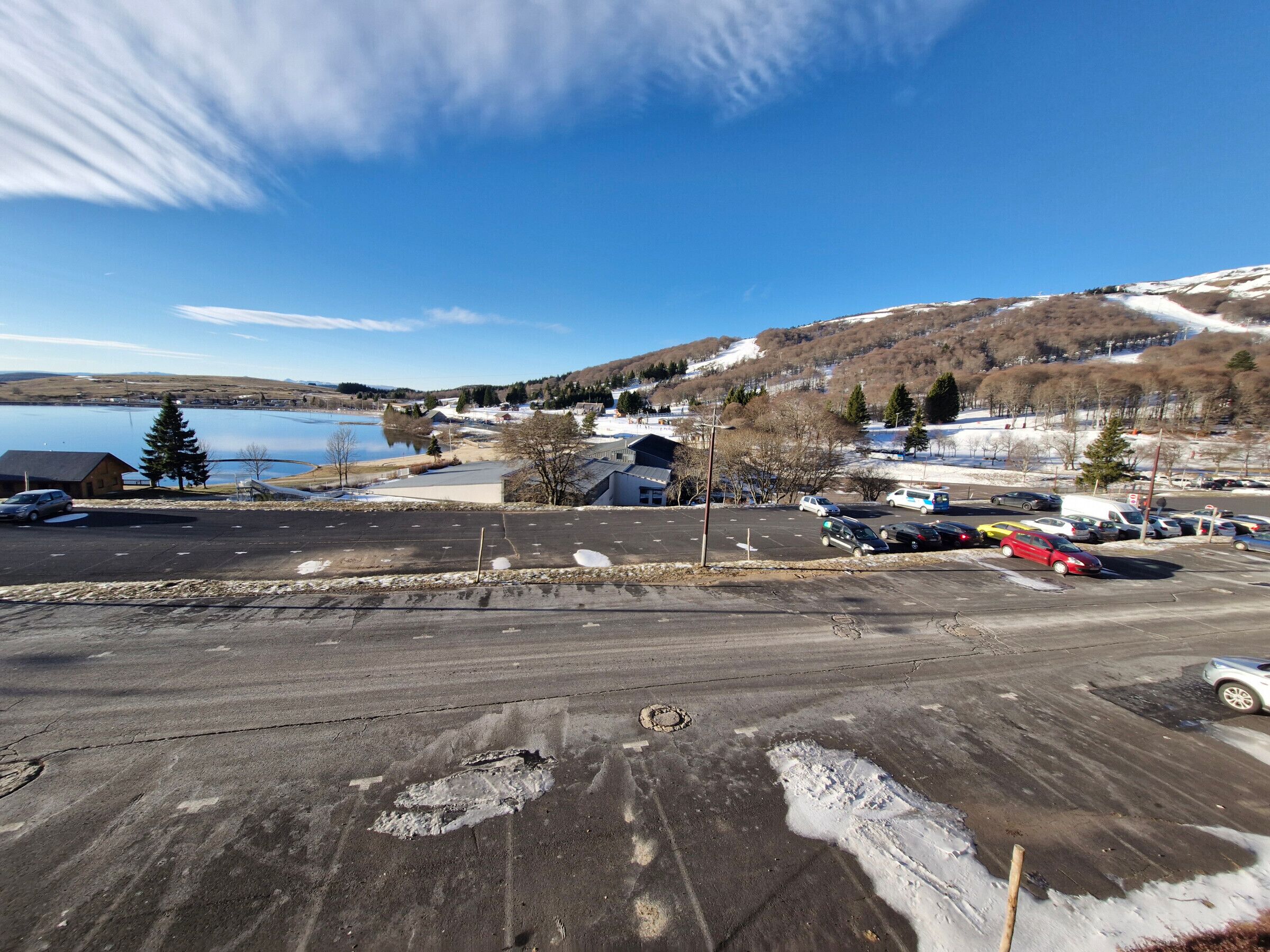 A parking lot with snow patches and a clear view of a lake and hills in the background.