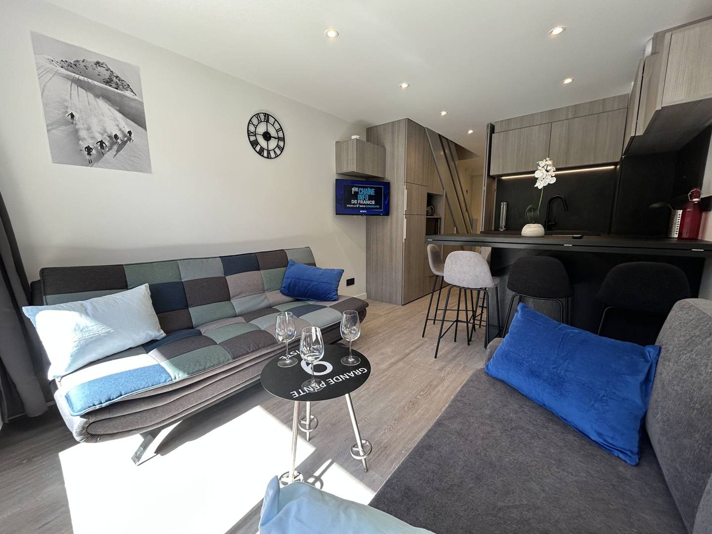 Een moderne woonkamer met een bank, een salontafel met glazen, een televisie, en een keuken met barstoelen.