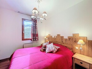 A bedroom with a wooden headboard, a large window with curtains, a chandelier, and bedside lamps. - Charming Renovated T3, South Balcony, Slope View (Le Corbier)