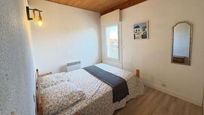 A small, cozy bedroom with a wooden ceiling, a bed with floral bedding, a bedside table, a window with a view, and a mirror on the wall. - Renovated 2023 house close to the ocean with terrace and WIFI (Biscarrosse Plage)