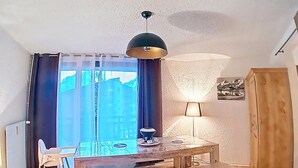 Zona de menjador amb taula i cadires de fusta, una làmpada penjant i vista cap a edificis a través de la finestra.
