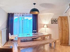 A dining area with a wooden table and chairs, a hanging lamp, and a view of buildings through the window. - Well-equipped studio at the foot of the slopes, central station (Les Deux Alpes)