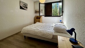Une chambre à coucher avec un lit, un bureau avec une lampe, une fenêtre donnant sur des arbres et une étagère fixée au mur.