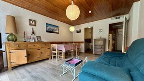 Un salon chaleureux avec un plafond en bois, un canapé bleu, une table à manger recouverte d’une nappe à carreaux et un buffet en bois doté de tiroirs.