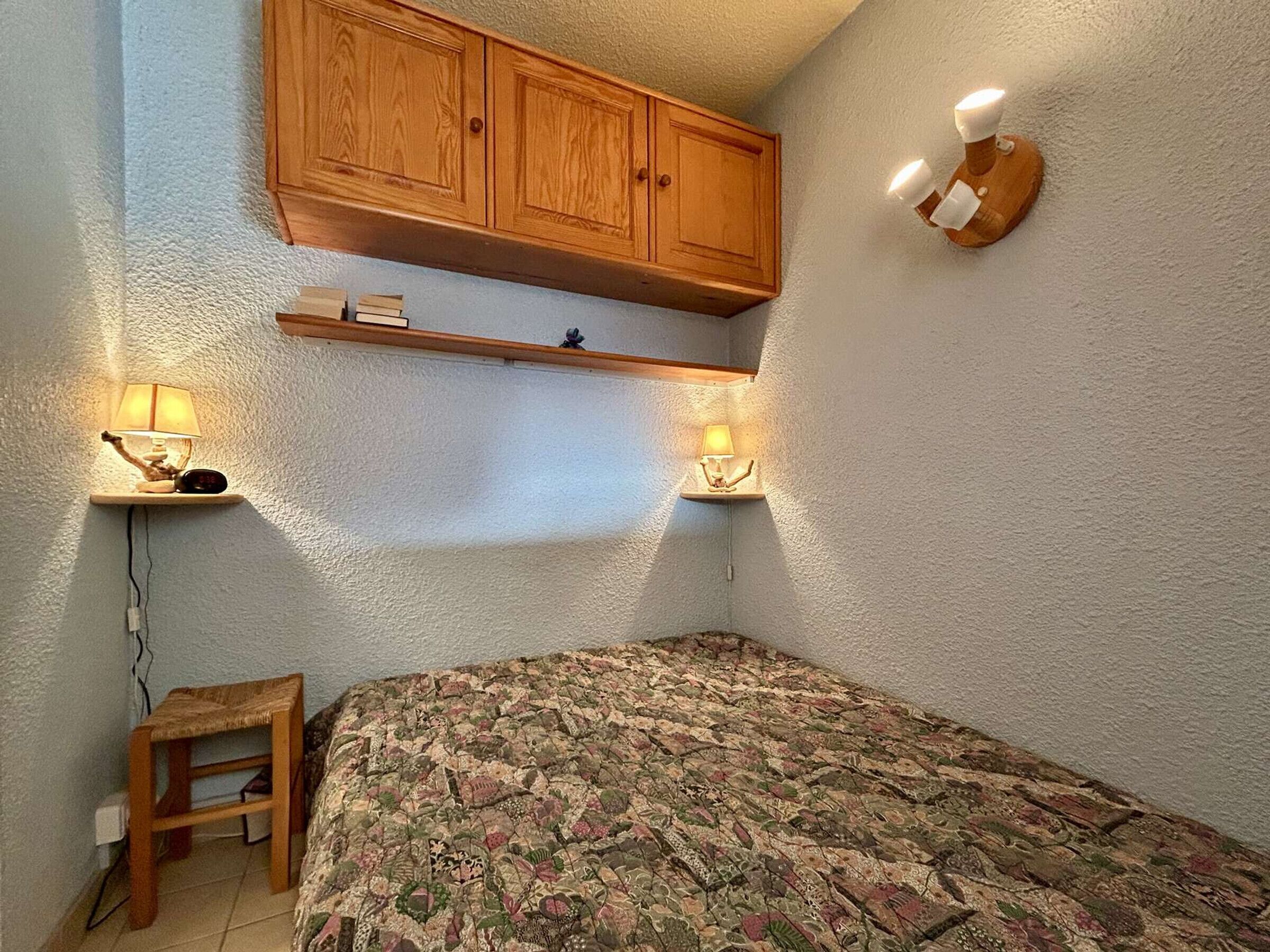 A bedroom with a wooden wardrobe, two wall-mounted lamps, a bed with a floral bedspread, and a small wooden stool.