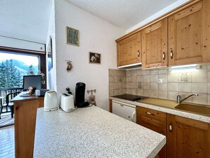A kitchen with wooden cabinets, a countertop, a stove, and a sink. There is a window with a view of trees and mountains. - Studio with balcony and parking - Briançon 4 people (Briançon)