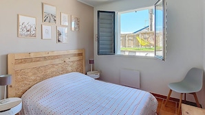A bedroom with a wooden headboard, a bed with a striped bedspread, a window with an open shutter, and a chair. - Villa in Vieux-Boucau for 10, near ocean, with terrace (Vieux-Boucau-les-Bains)