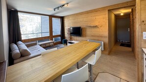 Een moderne keuken met een houten tafel en stoelen, een ingebouwde televisie en een groot raam met uitzicht op bomen.