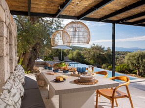Uma área de refeições ao ar livre com mesa de pedra, cadeiras e vista para uma piscina e paisagem.