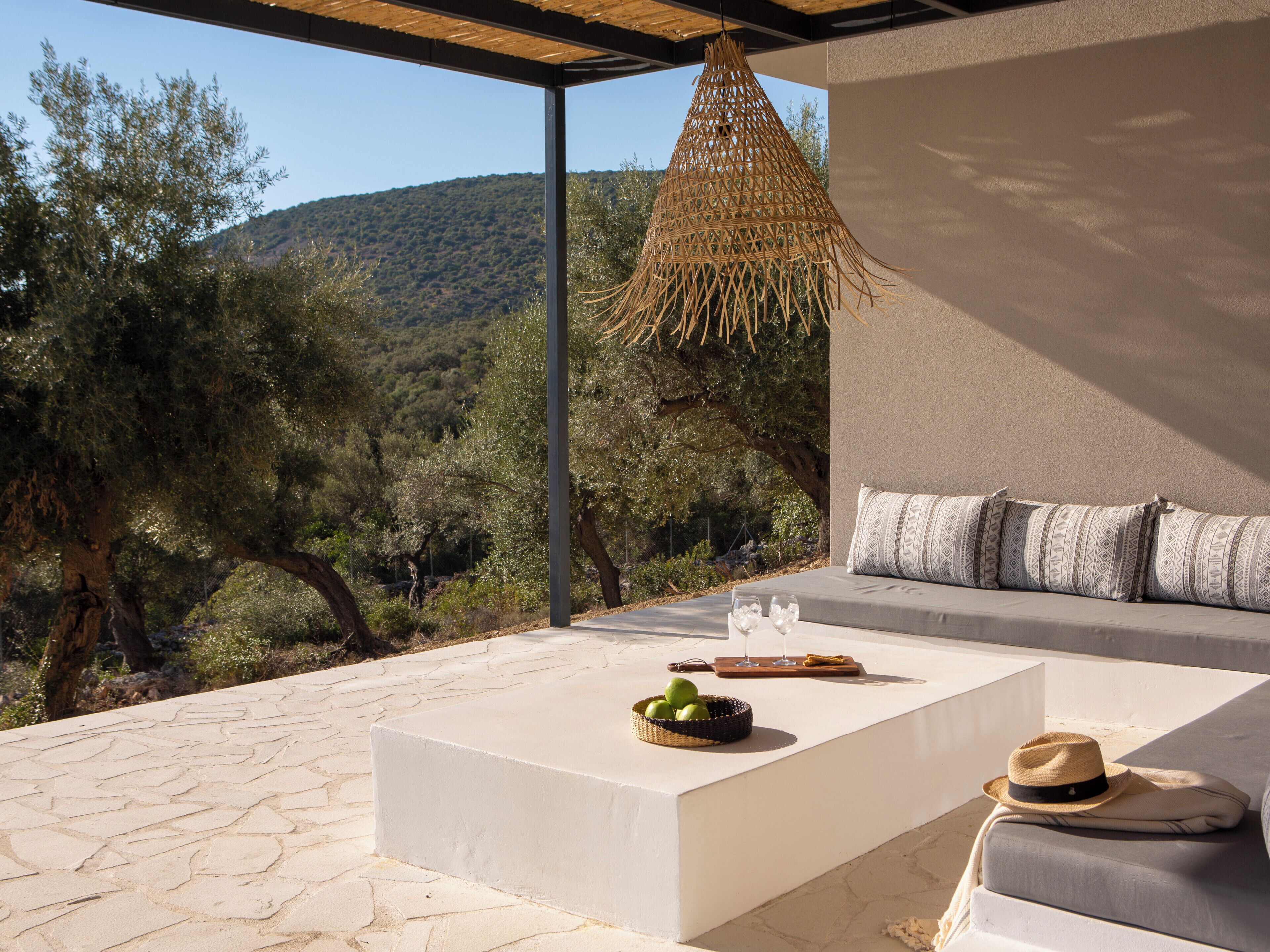 A modern outdoor seating area with a stone floor, a cushioned sofa with patterned cushions, a woven hanging light fixture, and a basket of green apples.