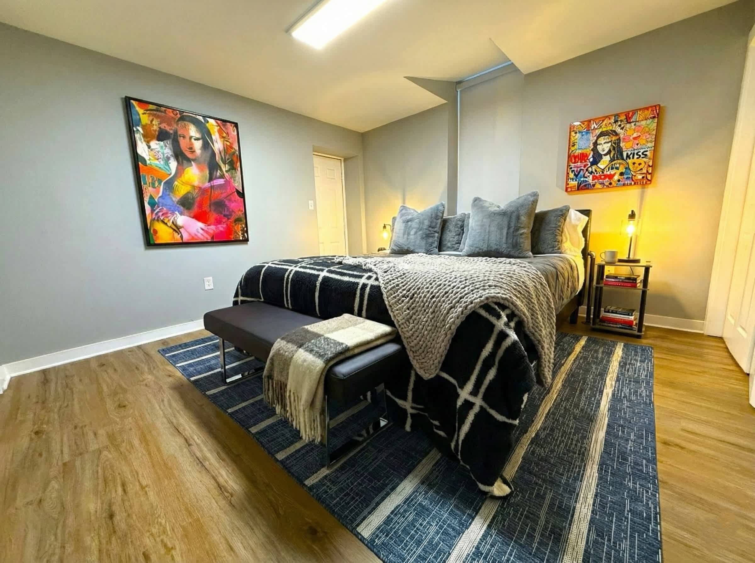 Une chambre à coucher comprenant un lit, un banc, une table de chevet, une lampe et deux tableaux accrochés aux murs.