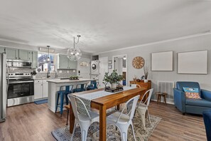 Eine moderne Küche mit Edelstahlgeräten, ein Essbereich mit einem Holztisch und weißen Stühlen sowie ein Wohnzimmer mit einem blauen Sofa und einer Wanduhr.