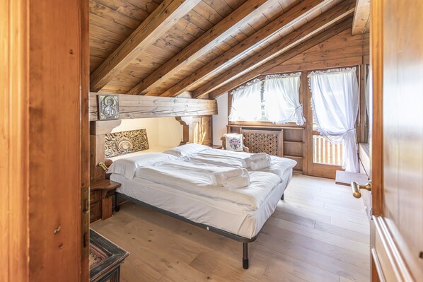 A wooden cabin bedroom with a bed, a nightstand, a chair, and a window with curtains. - Casa Lavinores, quiet and comfortable (Cortina d´Ampezzo)