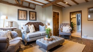 Uma sala de estar acolhedora com um sofá, poltronas e uma mesa de centro. Há quadros nas paredes e uma porta de madeira.