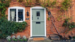 Uma casa de tijolos com porta verde, janela emoldurada em branco e um pequeno jardim com plantas e tapete de entrada.
