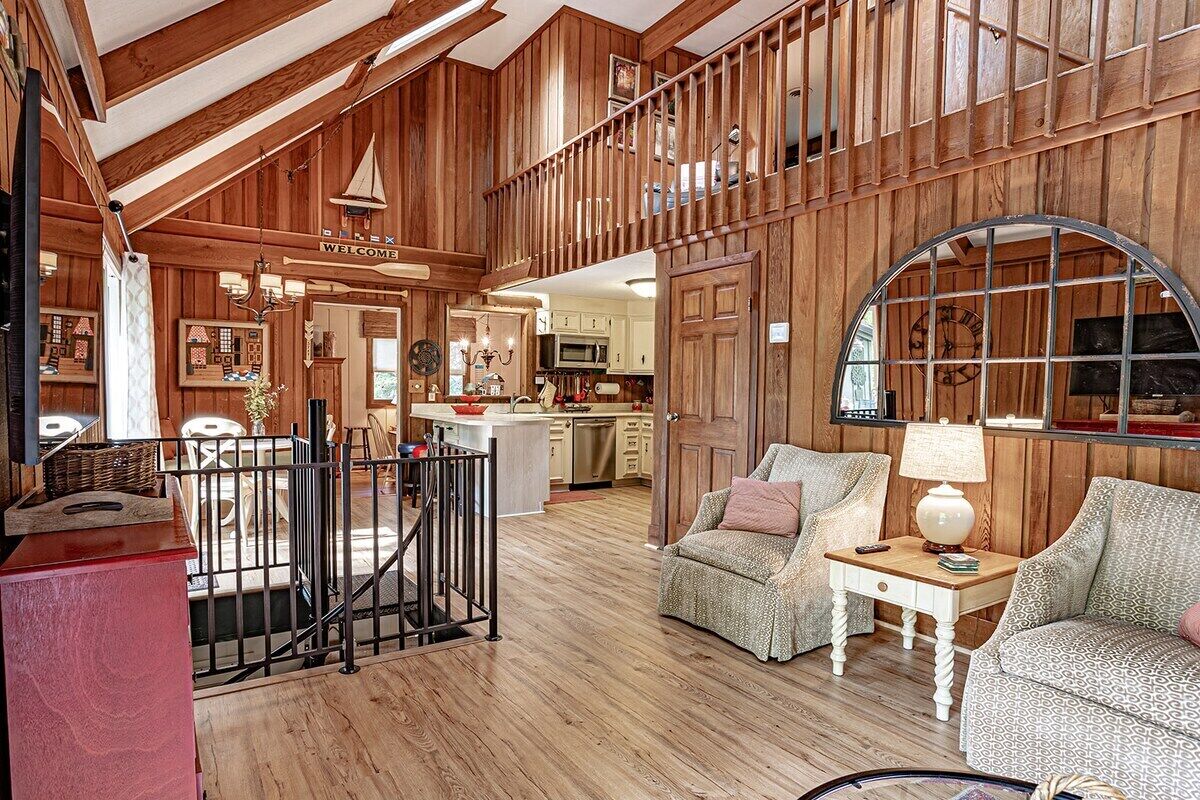 A spacious living area with a high ceiling, wooden walls, a staircase, and a kitchen in the background.