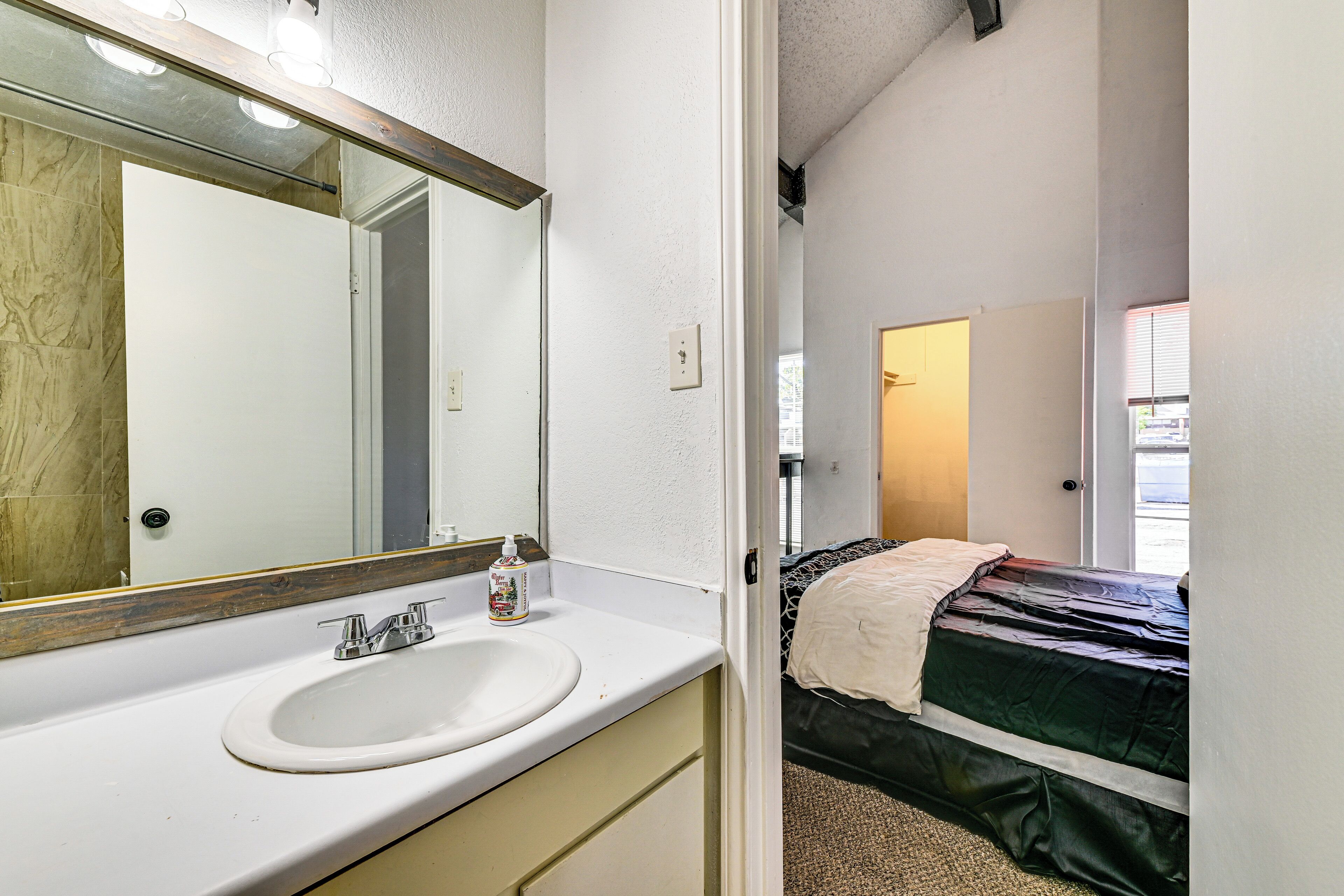 Un baño con lavamanos, espejo y una puerta que da a un dormitorio con cama y ventana.
