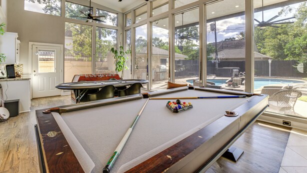 A pool table in a room with large windows, a ceiling fan, and a view of a backyard with a pool. - Heated Pool, Hot Tub, Game Room: Houston Group Gem (Houston)