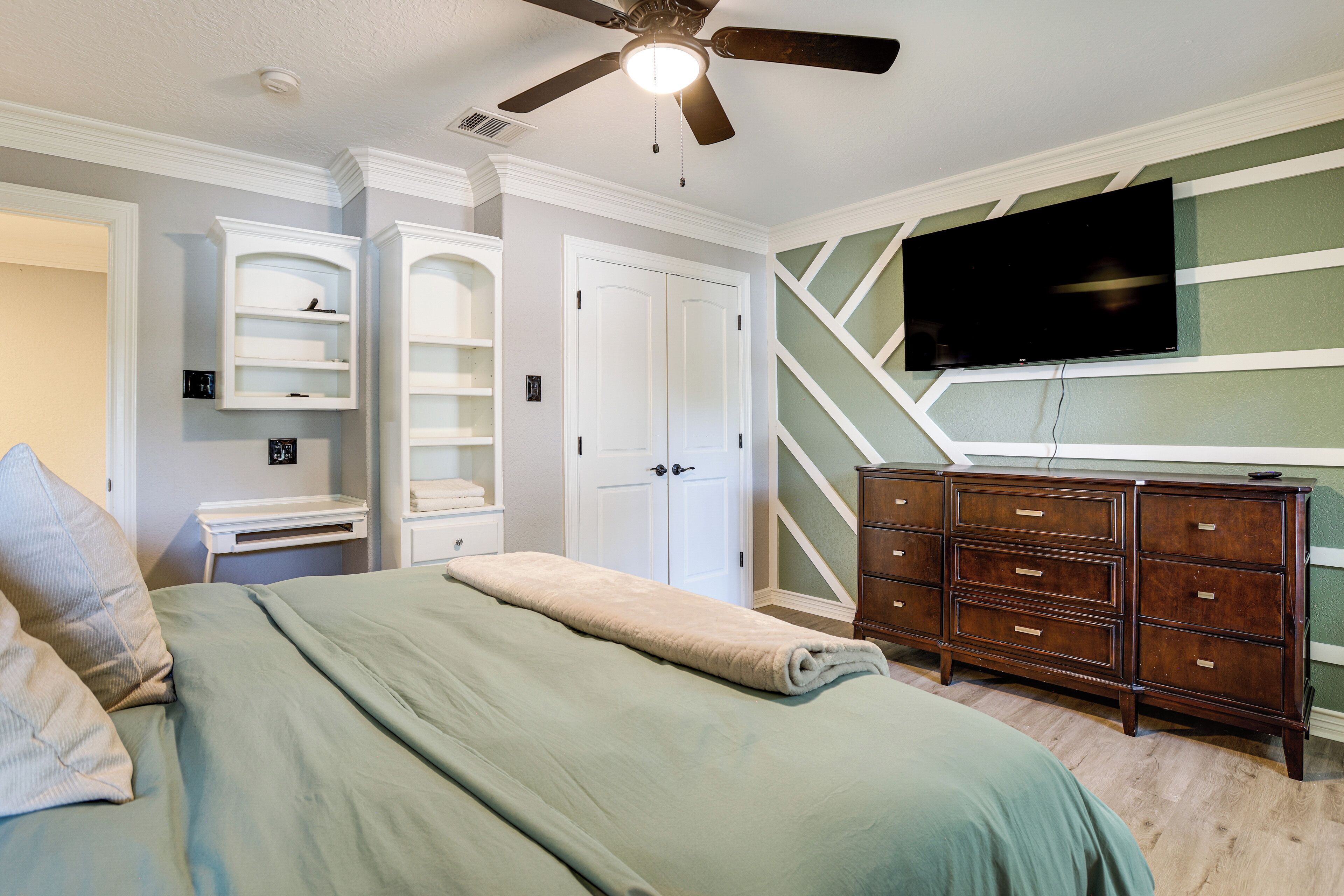 Une chambre à coucher comprenant un lit, un ventilateur de plafond, un téléviseur fixé au mur, une commode et des étagères intégrées.