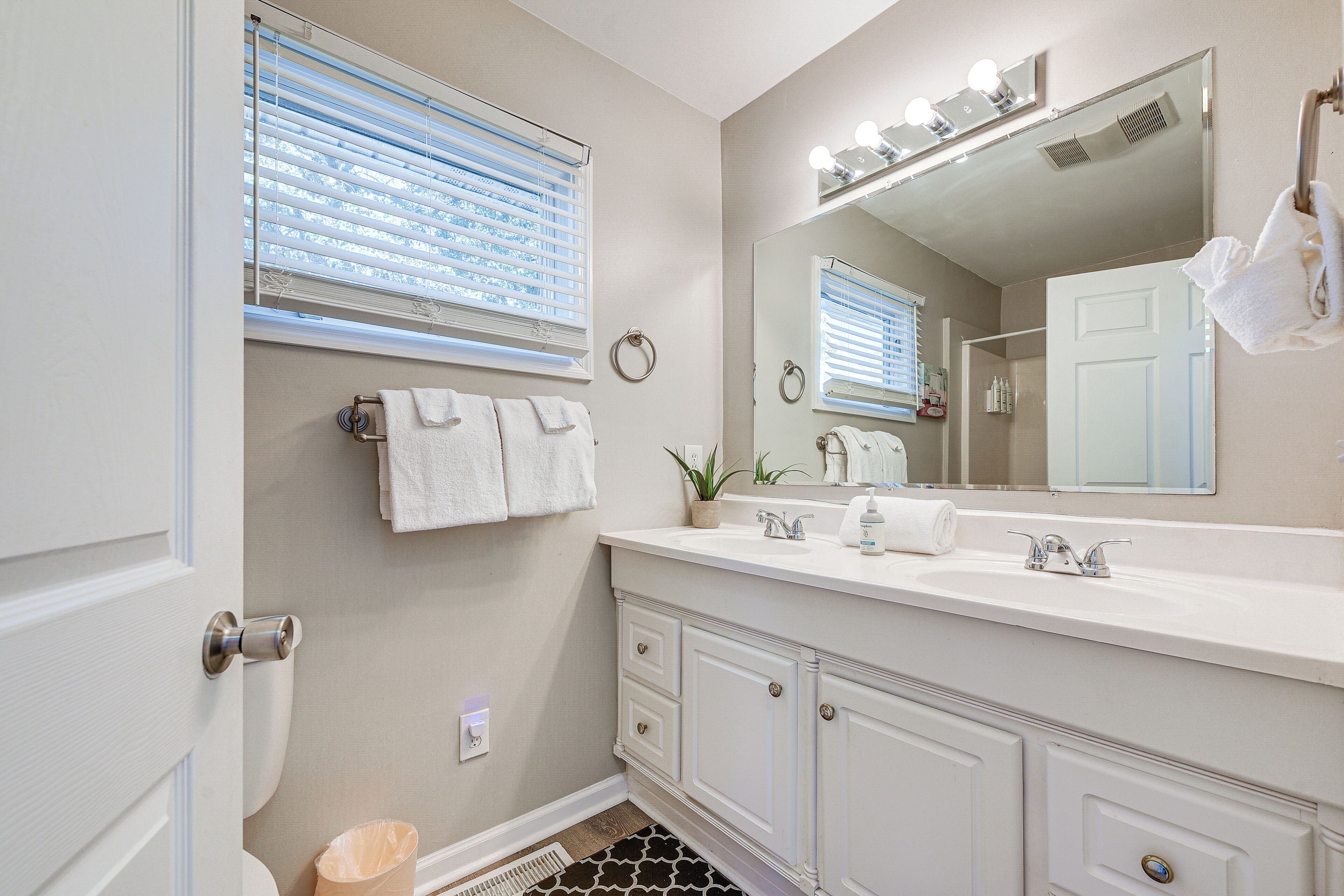 Une salle de bain avec un meuble-lavabo blanc, un grand miroir avec éclairage intégré, un porte-serviettes et une plante en pot.