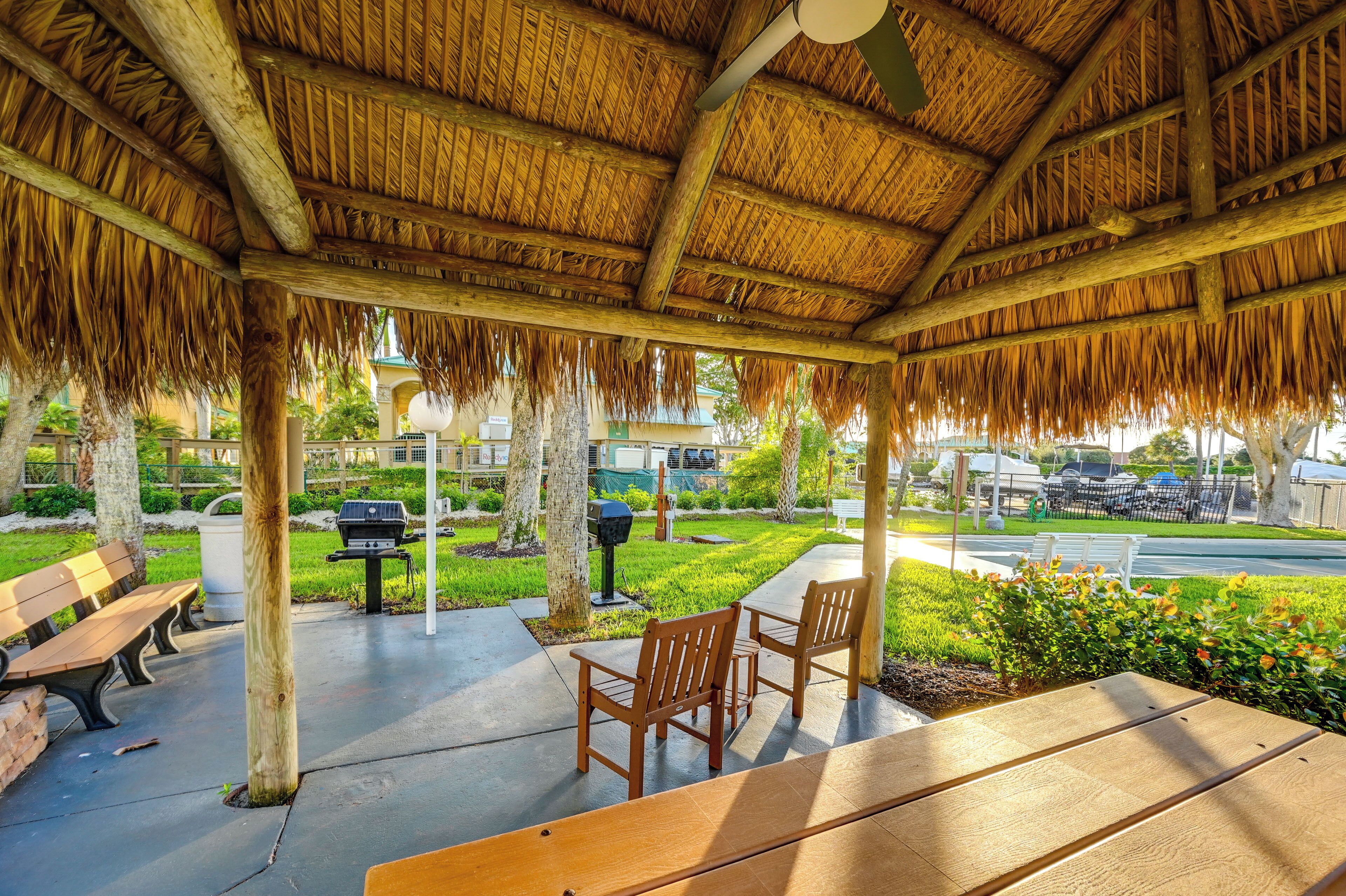 A covered outdoor seating area with wooden benches and chairs, a grill, and a view of a road and greenery.