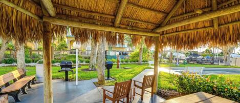 A covered outdoor seating area with wooden benches and chairs, a grill, and a view of a road and greenery.
