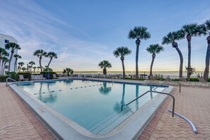 A rectangular pool with clear blue water, surrounded by palm trees and lounge chairs, with a view of the ocean in the background. - Riverside Marco Island Condo: Pool & Marina Access (Marco Island)